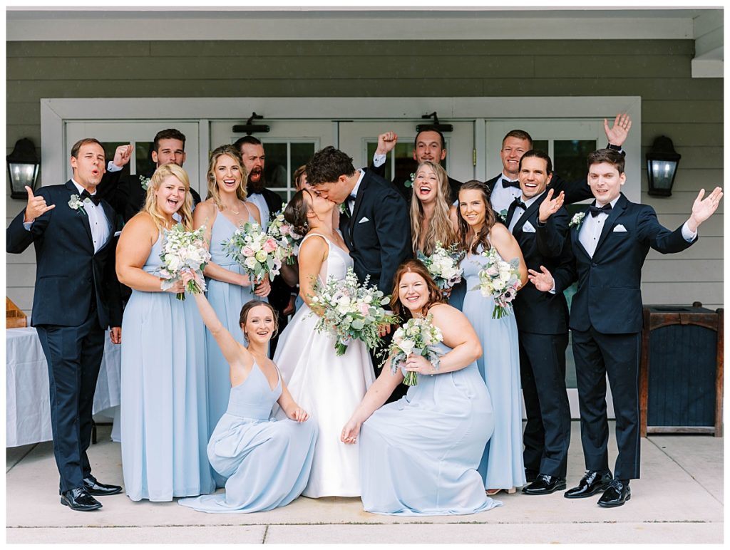 wedding party sharing a photo while bride and groom kiss at the chapel hill country club