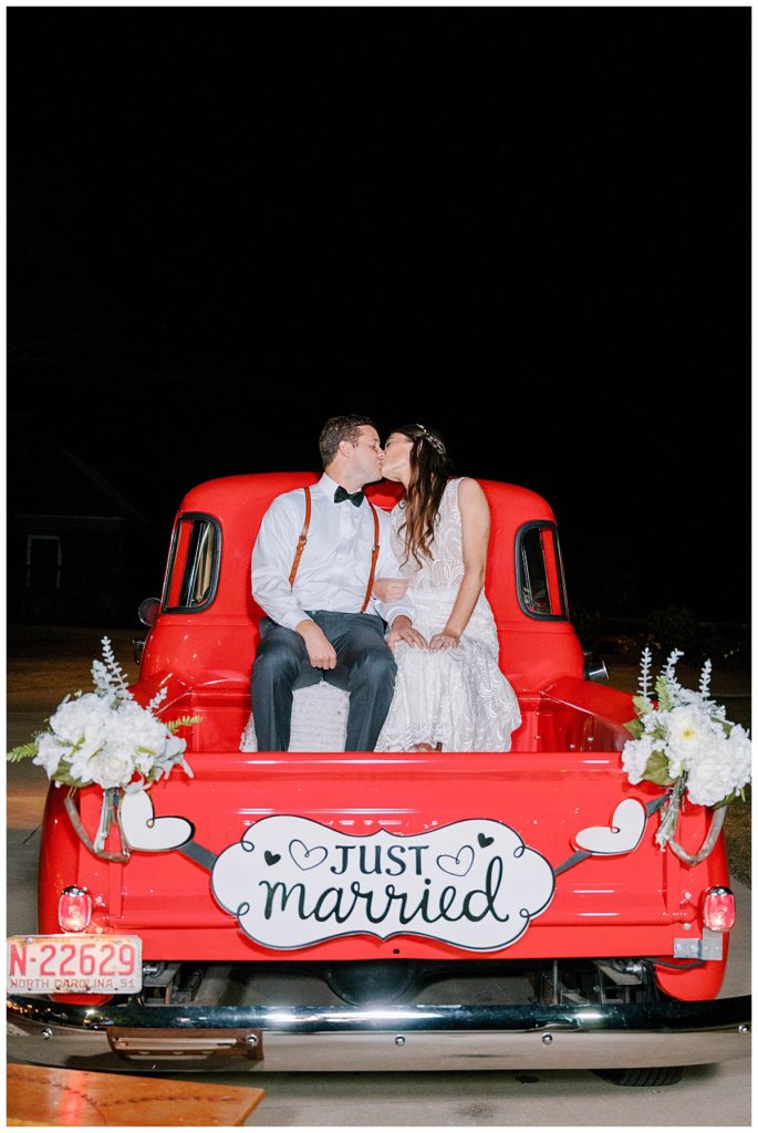 NEWLYWED BRIDE AND GROOM SITTING ON THE BACK OF RED CHEVY TRUCK THAT SAYS JUST MARRIED AT THE WALNUT HILL